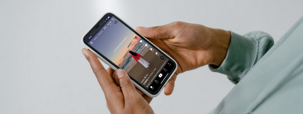 Close-up of a person holding a smartphone with both hands, displaying a TikTok video of a woman standing on a beach at sunset. The woman in the video is wearing a red shawl and white dress, facing the ocean. The TikTok interface shows typical engagement icons on the right, with the "For You" tab active at the top.