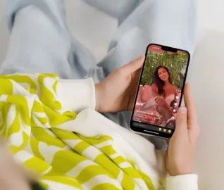 Person lounging casually while holding a smartphone, watching a TikTok video of a smiling woman in a light pink outfit seated on a couch with tropical plants in the background. The TikTok interface displays likes, comments, and caption text in a mix of emojis and script. The viewer is wearing light blue jeans and a lime green and white patterned sweatshirt.